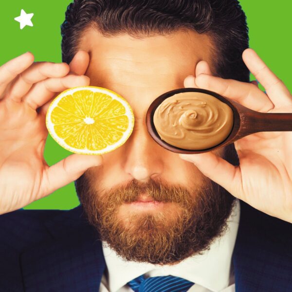 Man with a beard holding a lemon slice and a wooden spoon with a creamy substance in front of his face.